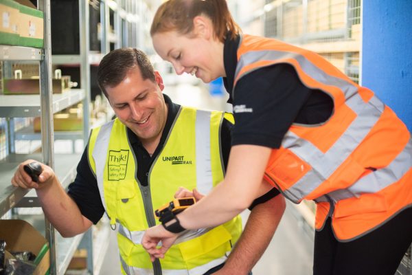 A woman in a Unipart Rail high visibility tabard speaking to a man on the apprenticeship programme in a warehouse.