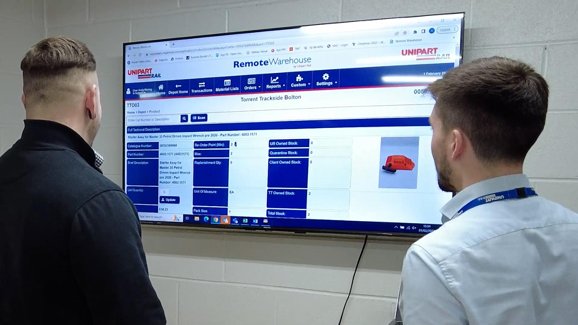 Two Unipart colleagues stood in front of a large wall mounted TV with remote warehouse software open showing details of the Torrent Trackside Bolton.