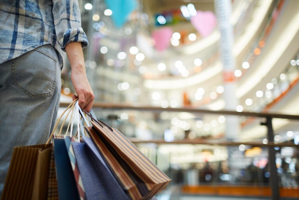 Close-up of woman in casual clothing standing in spacious shopping mall with lights and holding many paper bags.