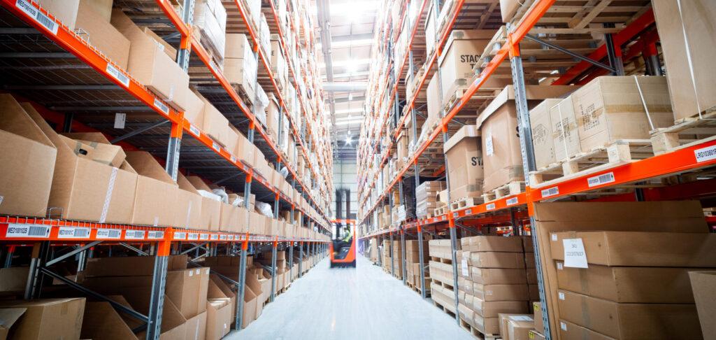 A Unipart logistics warehouse showing boxes stacked on metal storage racks and a forklift visible in the distance in the aisle.