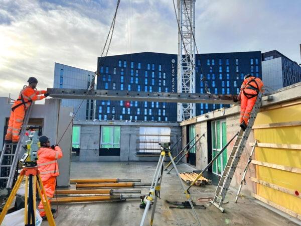 Construction workers in high-visibility orange safety gear are assembling a steel beam on a building site.