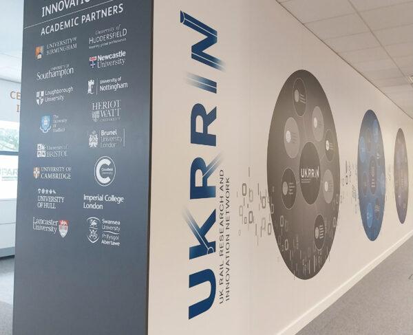 Wall at the UKRRIN technology hub showing UKRRIN logo and logos of academic partners.