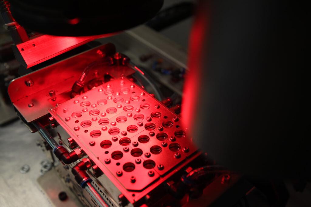 A close-up view of a precision-engineered metal fixture with multiple circular holes, illuminated by a red inspection light. Part of a manufacturing quality control process, with various mechanical and pneumatic components visible.
