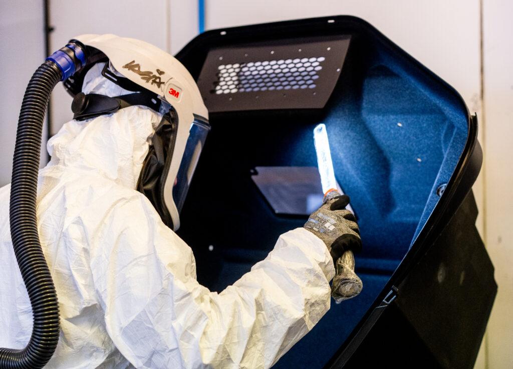 A worker wearing a full protective suit, gloves, and a powered air-purifying respirator (PAPR) helmet with a hose is performing a task inside a specialized workstation.