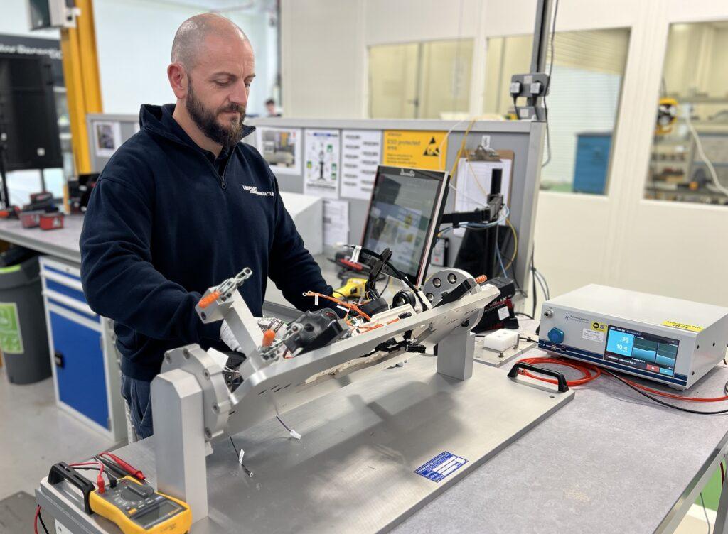 A technician in a navy-blue uniform is working on an assembly fixture in a laboratory or manufacturing environment. He is handling a complex mechanical and electrical component, which includes wiring and connectors. A computer monitor displaying technical data is mounted on a stand in front of him, and various testing equipment, including a multimeter and an electronic testing device, are on the workbench.