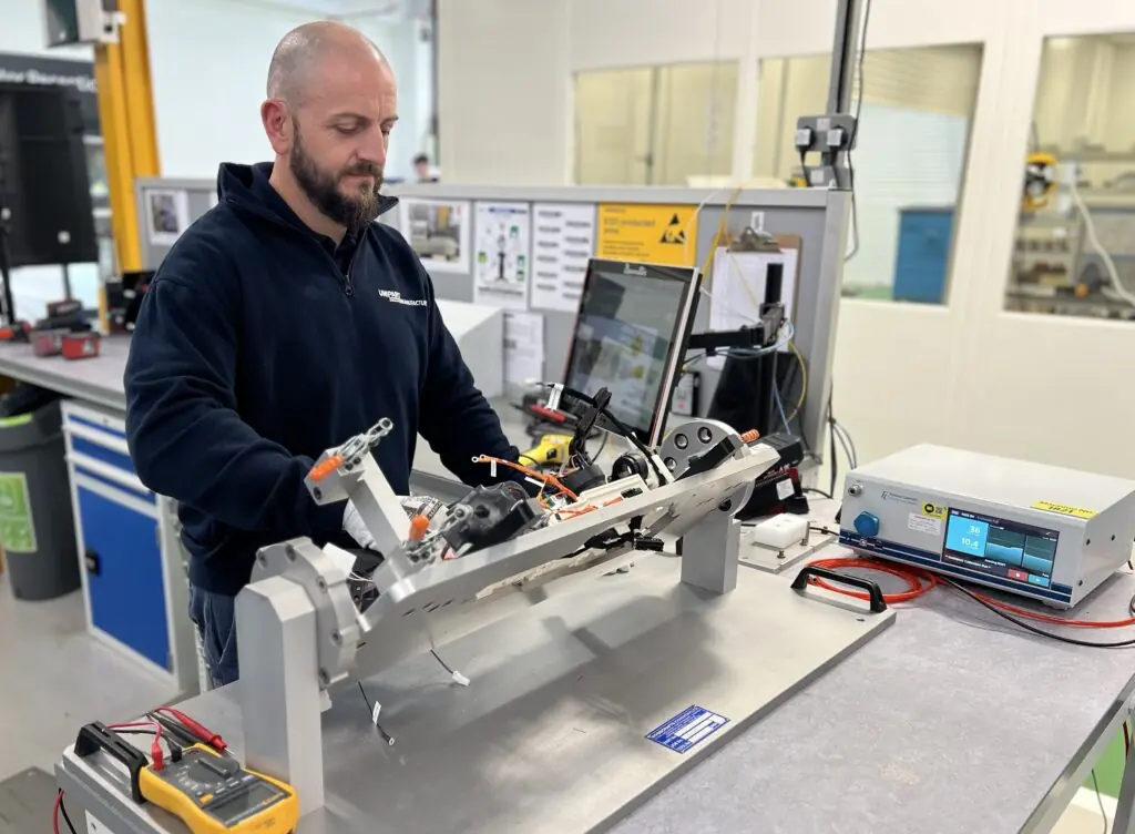 A technician in a navy-blue uniform is working on an assembly fixture in a laboratory or manufacturing environment. He is handling a complex mechanical and electrical component, which includes wiring and connectors. A computer monitor displaying technical data is mounted on a stand in front of him, and various testing equipment, including a multimeter and an electronic testing device, are on the workbench.